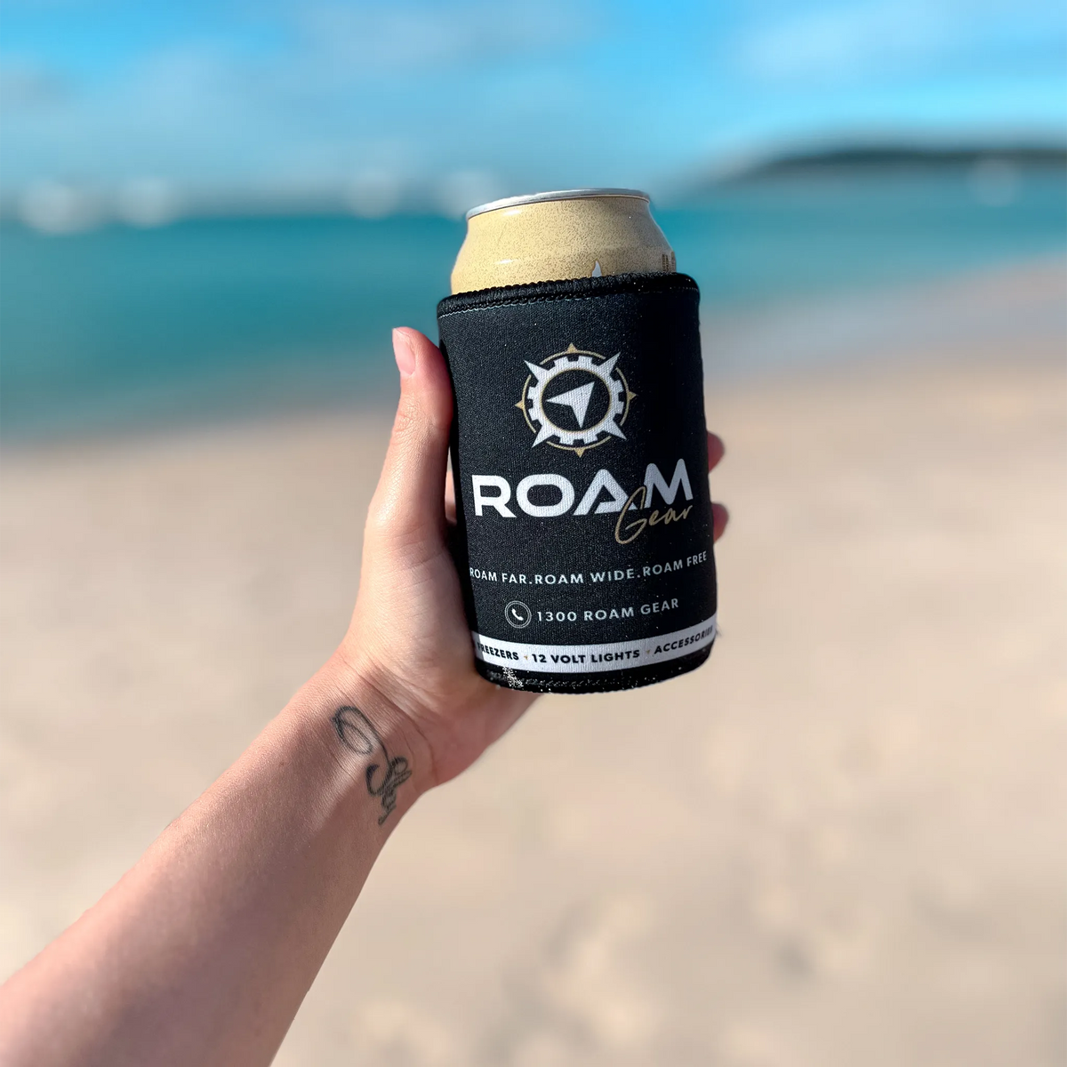 Roam Gear black stubby cooler holding a can in a person's hand with the beach and ocean behind.