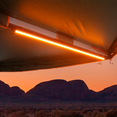 Hardkorr 100cm LED light bar mounted beneath an awning, glowing orange with mountains beyond at sunset.