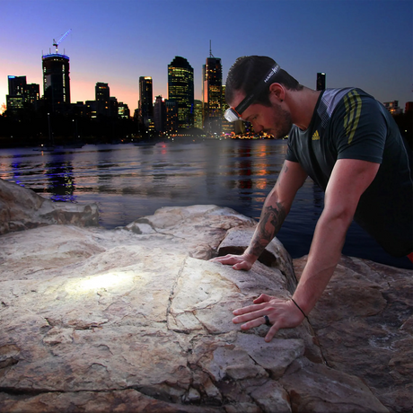Hardkorr 145 lumen headtorch lighting a rock beside the water with city buildings in the background.
