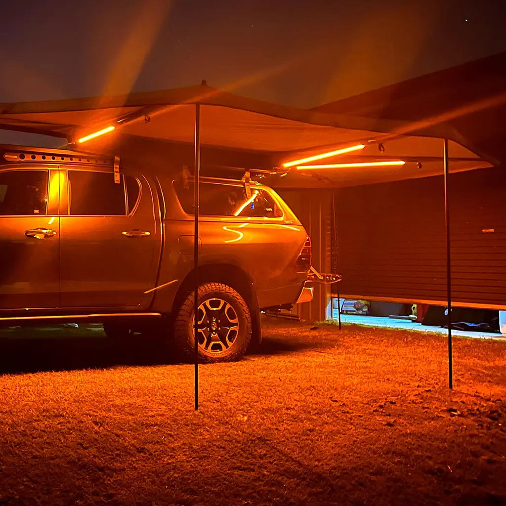 Hardkorr 3 strip LED awning light kit casting orange light beneath a vehicle awning at night.