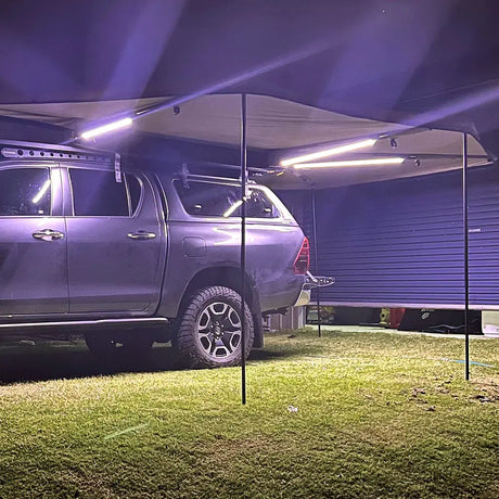 Hardkorr 3 strip LED awning light kit casting white light over the grass beside a vehicle.