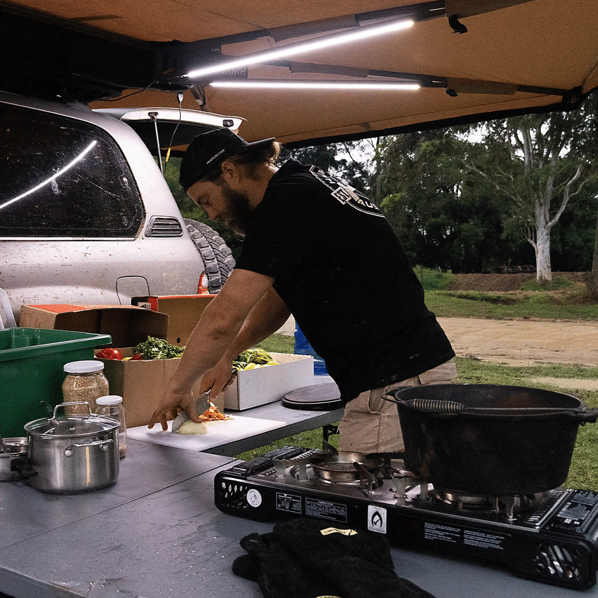 Hardkorr 3 strip LED awning light kit lighting a camp kitchen while a man prepares food.