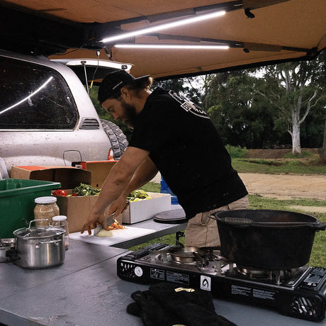 Hardkorr 3 strip LED awning light kit lighting a camp kitchen while a man prepares food.