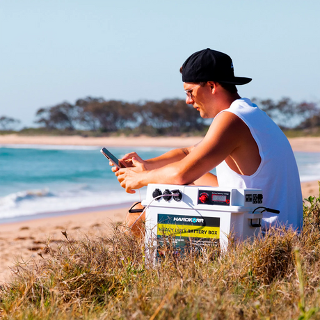 White Hardkorr battery box beside a person using a phone on a grassy sand dune near the beach.