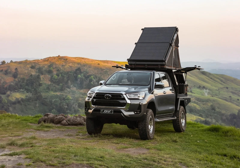 Toyota HiLux with an open hard-shell rooftop tent on a grassy hilltop