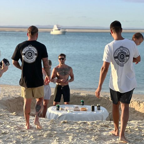 Two men wearing the Roam Gear "Live the Roam Life" T-shirt from the rear at a beach gathering.