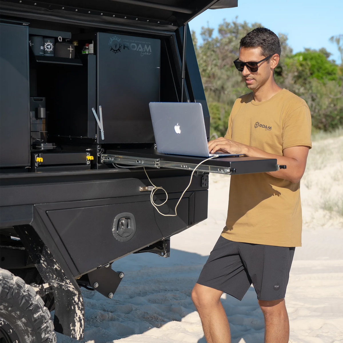Man wearing the Roam Gear "Live the Roam Life" male tan T-shirt while using a laptop beside a vehicle canopy.