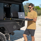 Man wearing the Roam Gear "Live the Roam Life" male tan T-shirt while using a laptop beside a vehicle canopy.