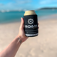Roam Gear black stubby cooler holding a can in a person's hand with the beach and ocean behind.